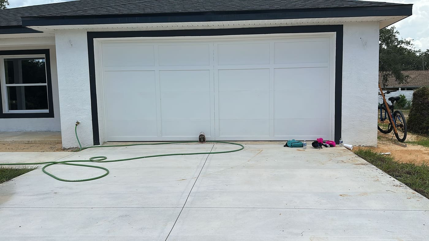 A white garage door with a dark surround on a newly finished house, seen across a clean concrete driveway with a hose on it.
