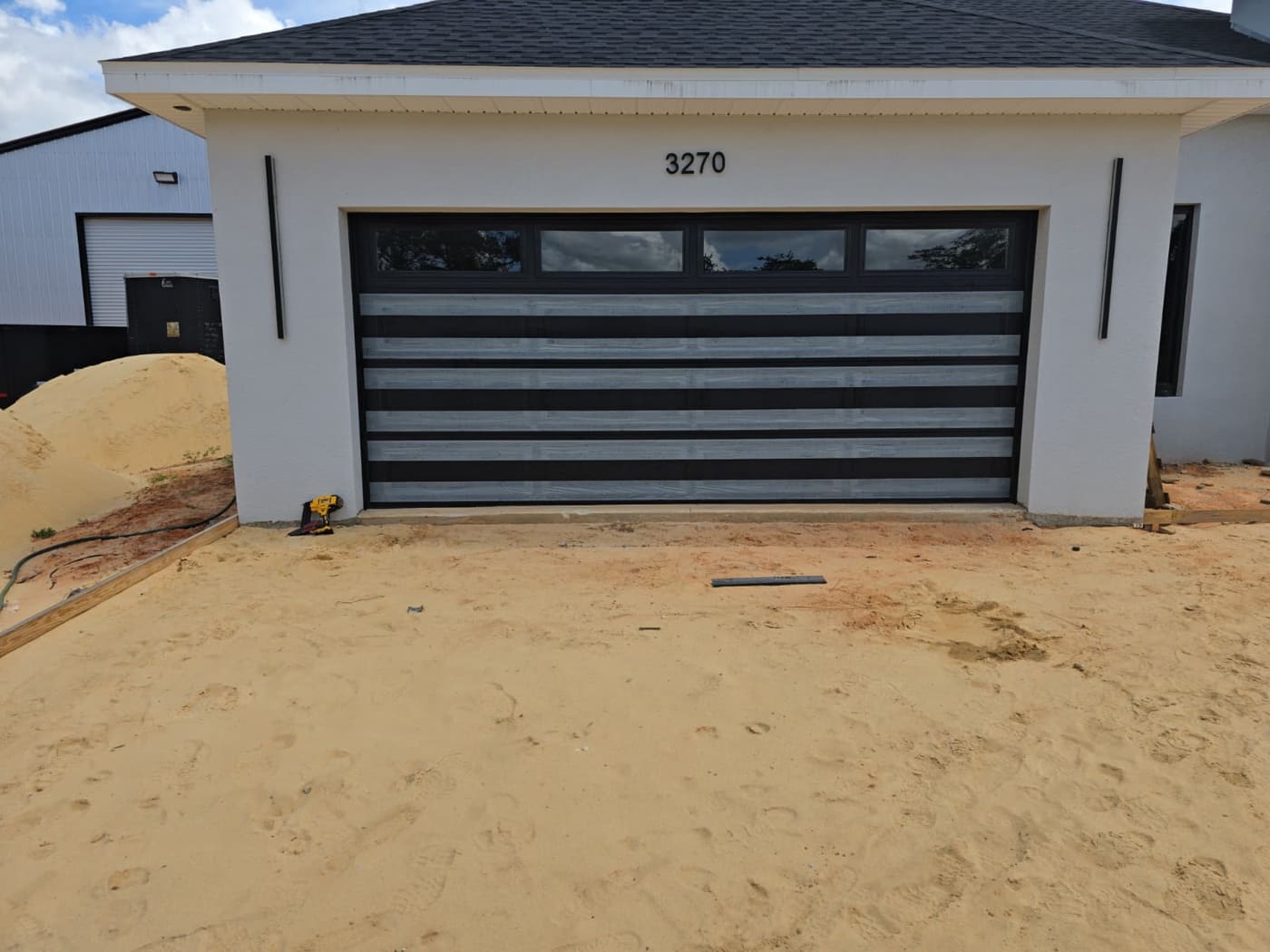 A wide double garage door in charcoal with pale horizontal bands and a row of tinted windows along the top, on a newly built white stucco house with the driveway still bare sand.