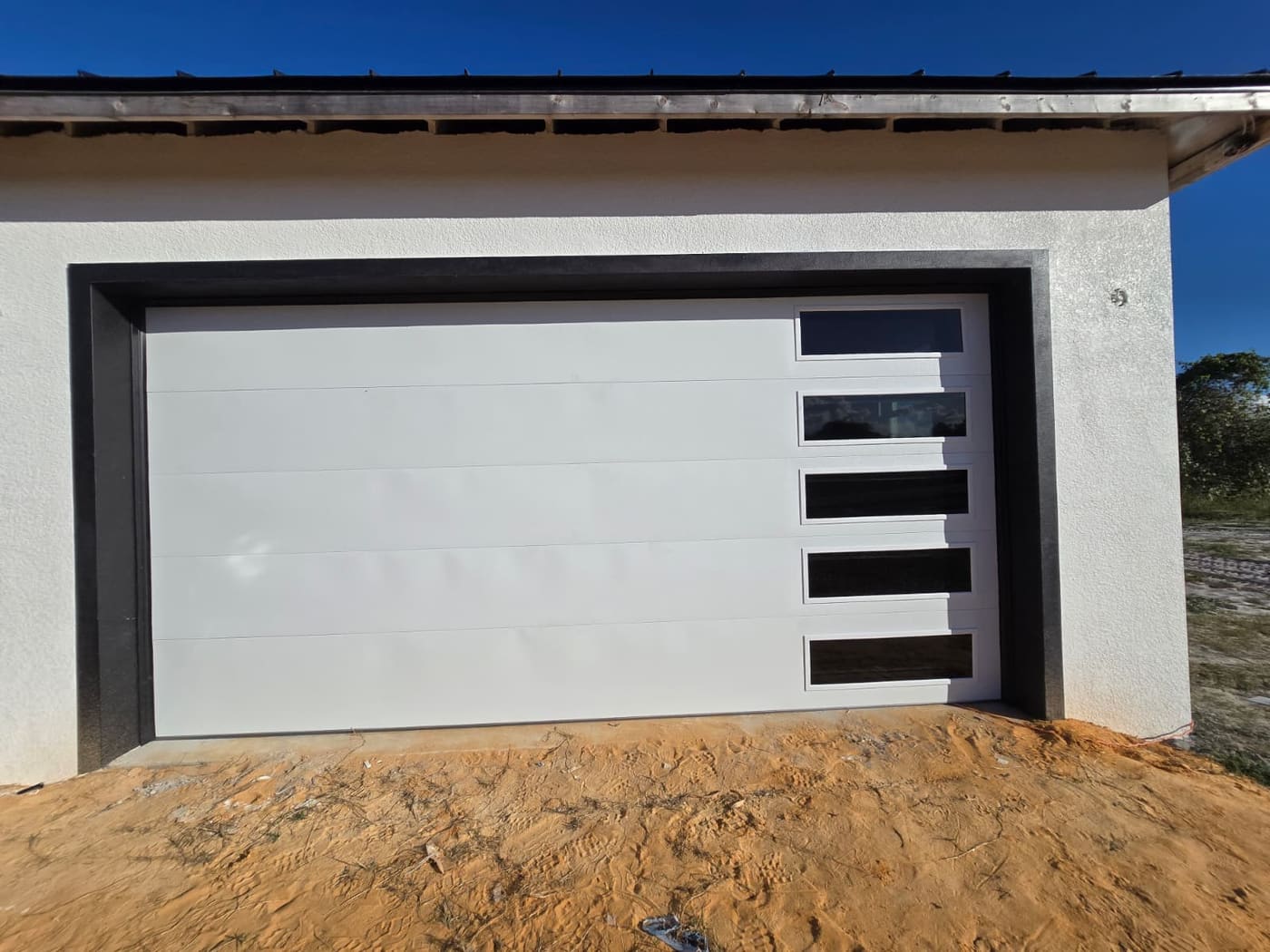 A flush white garage door with a vertical row of narrow windows, on a house whose driveway is still sand.