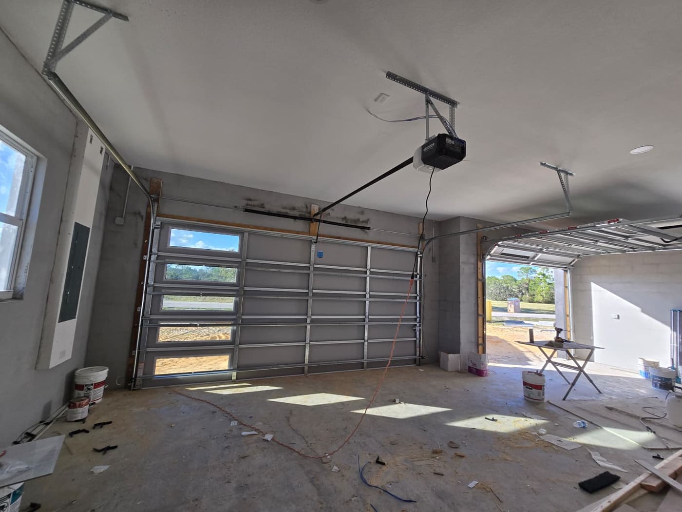 The inside of a two-bay garage under construction. One sectional door is hung with its track curving up to the ceiling and an opener on a rail; the second opening is still bare, showing the track fitted and no door on it.