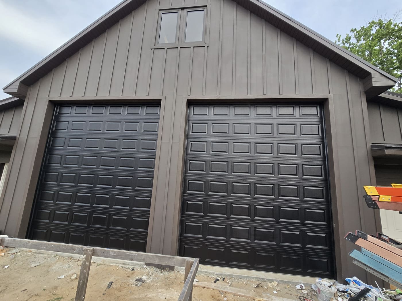 Two black garage doors side by side on a tall barn-style building clad in dark board, with building materials stacked in front.