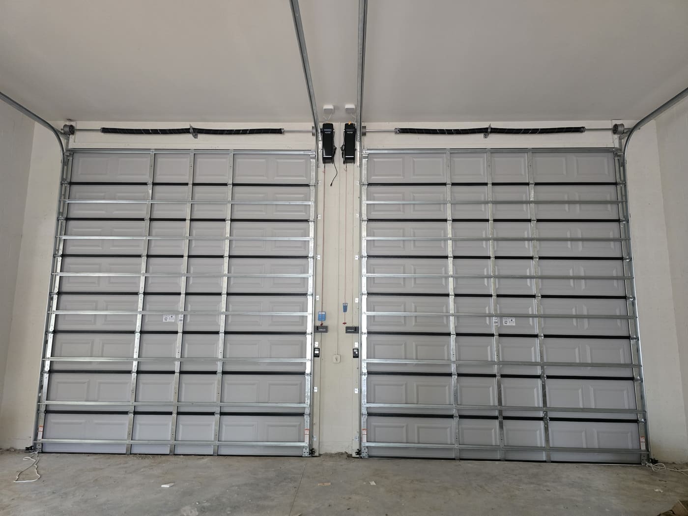 Two grey raised-panel garage doors side by side seen from inside a garage, each with steel struts across the panels, its own torsion spring on a shaft above the opening, curved track running back to the ceiling and a wall control beside it.