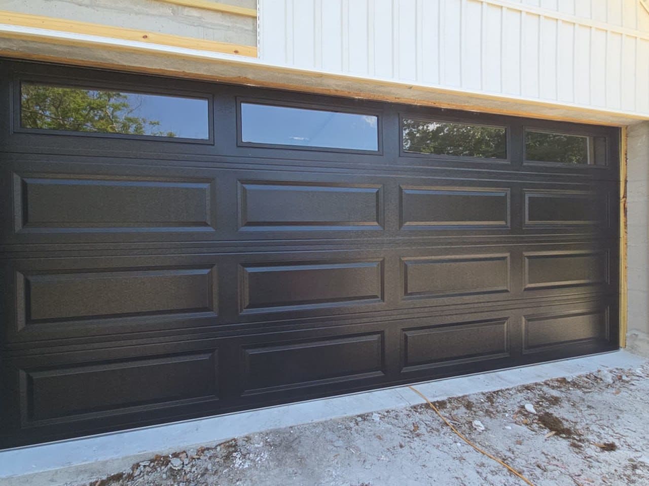 A dark brown long-panel double garage door with four windows in the top section, on a house with a fresh concrete apron.
